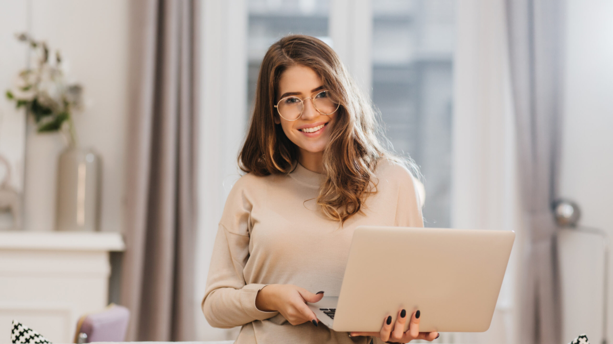 Elegant female student in glasses posing with pleasure, holding laptop. Refined brunette girl in beige shirt working with computer in her room.