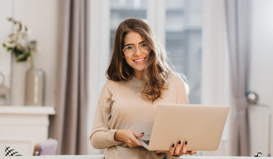 Elegant female student in glasses posing with pleasure, holding laptop. Refined brunette girl in beige shirt working with computer in her room.